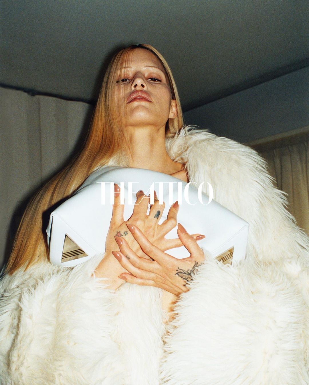 Adult woman poses with a large, white purse and fur coat. The image is taken from a low angle, highlighting fashion elements.