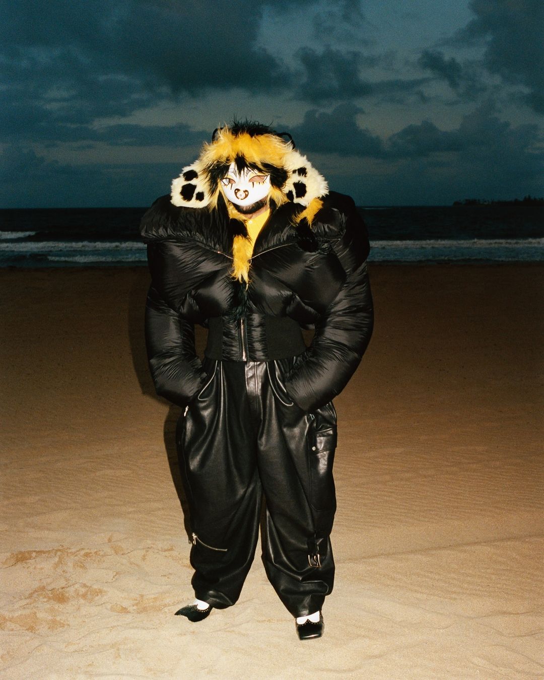 A person in a black puffy jacket and pants stands on a sandy beach under a cloudy sky, creating a unique fashion portrait.
