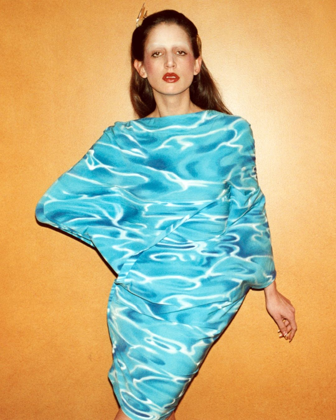 A fashion shot of a slim woman with fair skin and light brown hair, wearing a turquoise dress with an aquatic pattern against a plain orange background.
