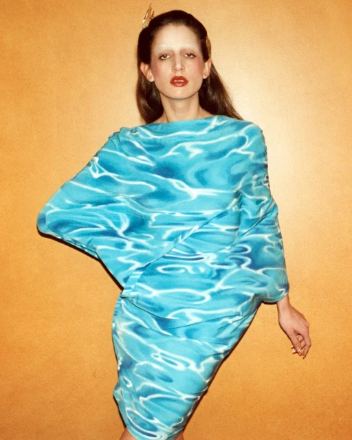 A fashion shot of a slim woman with fair skin and light brown hair, wearing a turquoise dress with an aquatic pattern against a plain orange background.