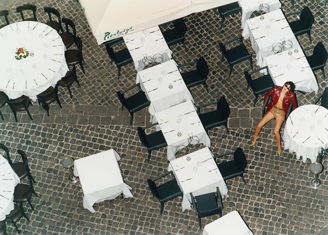 Overhead view of an adult woman in a red jacket lying on cobblestones among white-tableclothed tables and chairs at an outdoor restaurant.