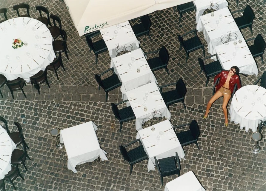 Overhead view of an adult woman in a red jacket lying on cobblestones among white-tableclothed tables and chairs at an outdoor restaurant.