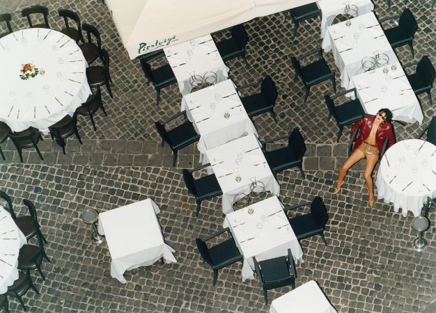Overhead view of an adult woman in a red jacket lying on cobblestones among white-tableclothed tables and chairs at an outdoor restaurant.
