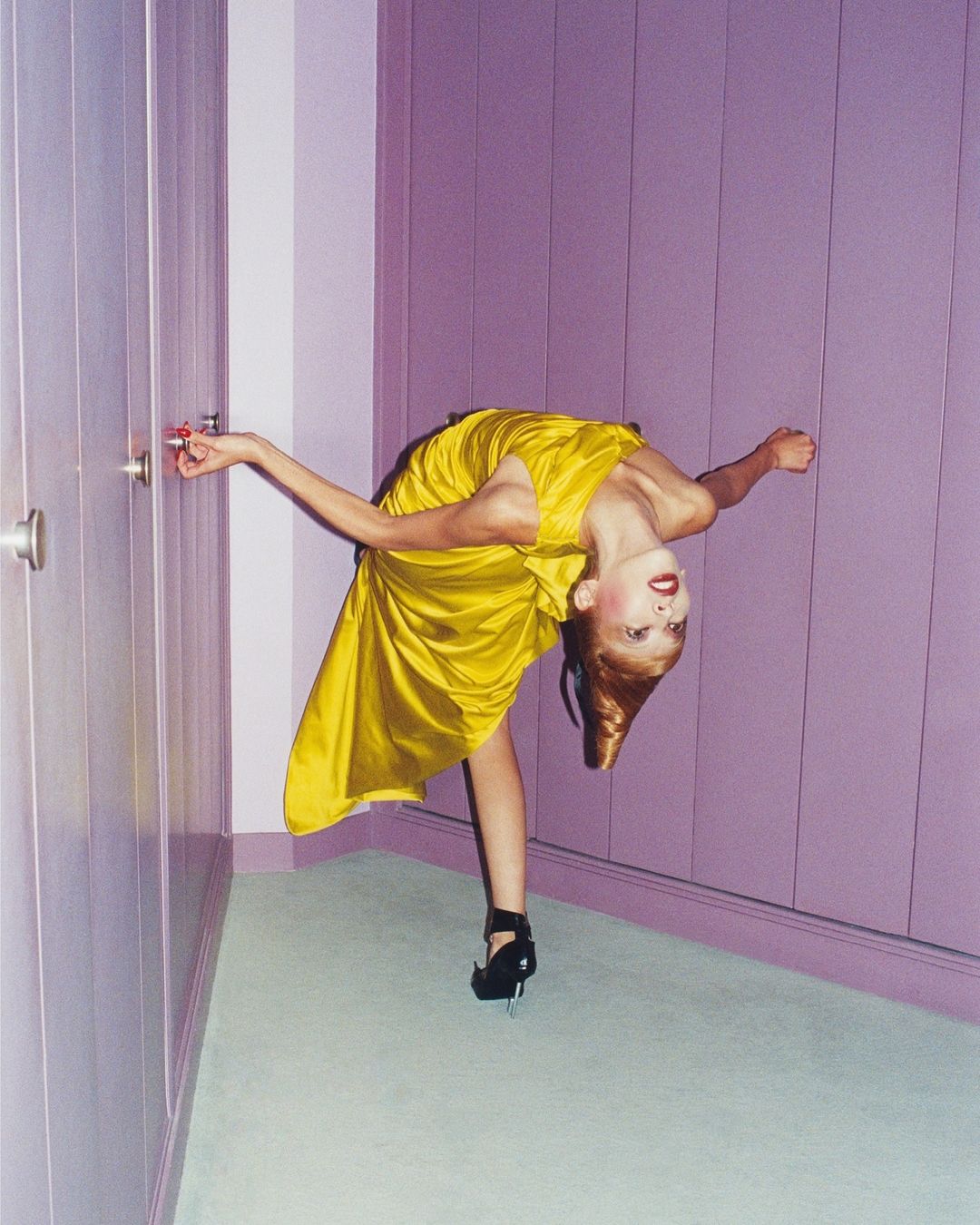 Red-haired woman in a yellow dress bends backward in front of a wardrobe. She wears black high heels and has fair skin and red lipstick.