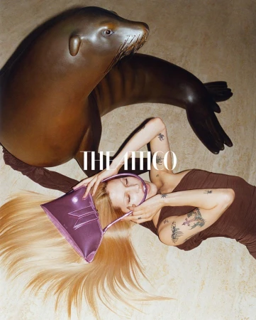 Fashion shot featuring a woman with blonde hair, light makeup, and a brown dress, lying next to a brown sea lion statue and purse.