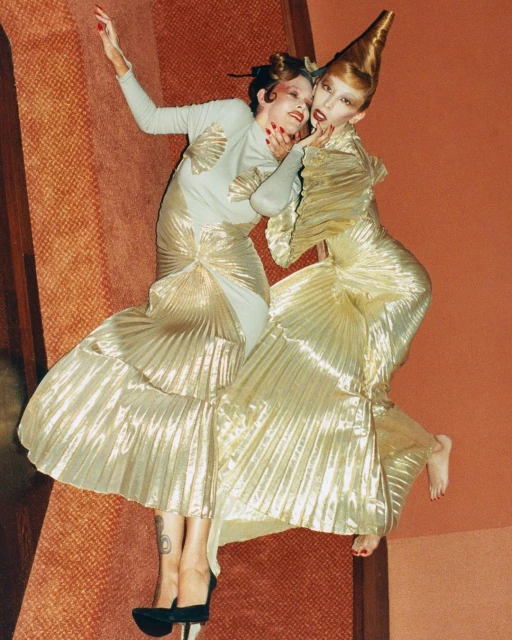 Two fashion models in avant-garde gold gowns pose on a rust-colored floor. One wears a cone-shaped headpiece and bold makeup.