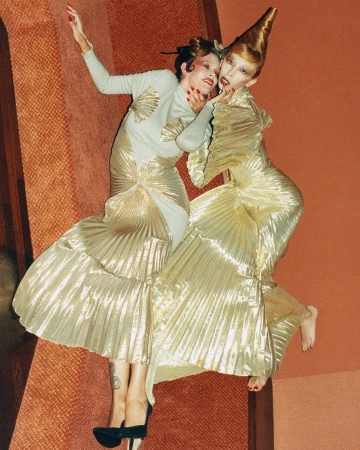 Two fashion models in avant-garde gold gowns pose on a rust-colored floor. One wears a cone-shaped headpiece and bold makeup.