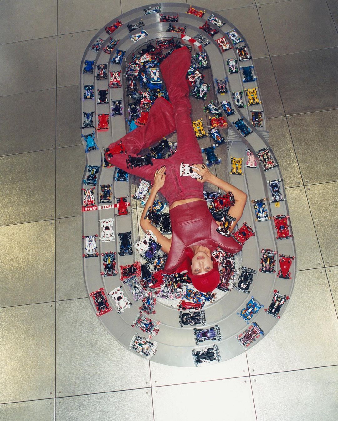 Overhead shot of an adult female lying on a toy race track surrounded by toy cars, wearing red pants, top, and boots on a tile floor.