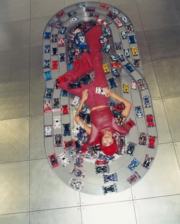Overhead shot of an adult female lying on a toy race track surrounded by toy cars, wearing red pants, top, and boots on a tile floor.