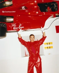 Model in red jumpsuit lifting a hanging red Formula One racing car wearing white gloves and glasses
