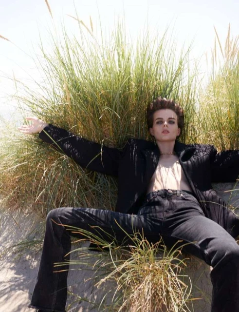 Woman in black blazer and jeans reclining in dune grass on sand in bright daylight in a fashion shot with casual, confident pose.
