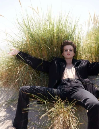 Woman in black blazer and jeans reclining in dune grass on sand in bright daylight in a fashion shot with casual, confident pose.