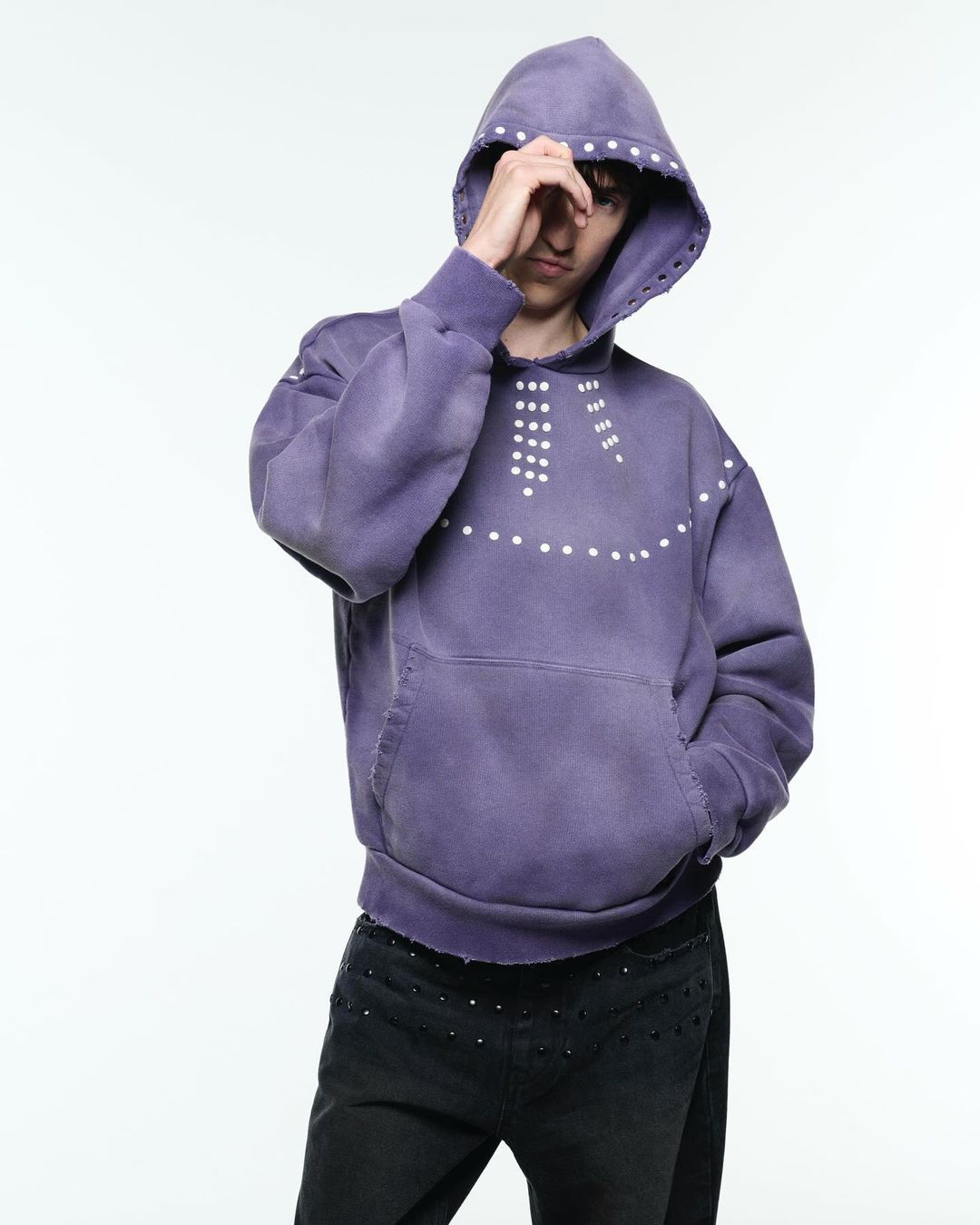 Adult male model poses in a lilac hooded sweatshirt with white dots, paired with black studded pants, against a bright white backdrop.