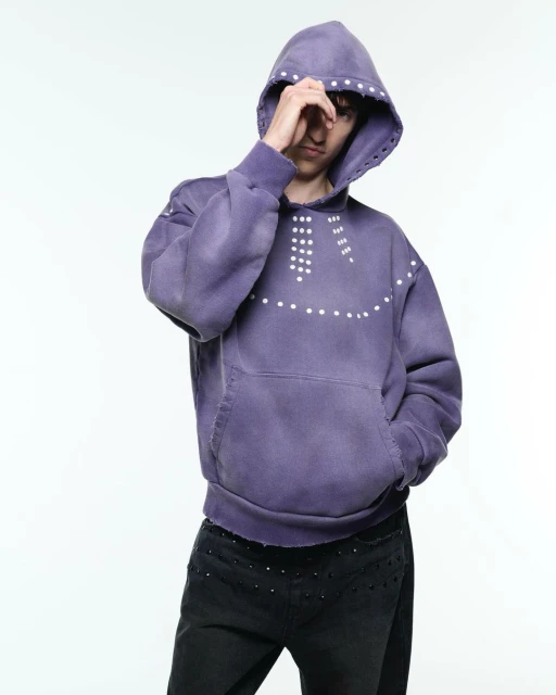 Adult male model poses in a lilac hooded sweatshirt with white dots, paired with black studded pants, against a bright white backdrop.