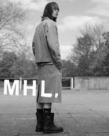 Monochrome fashion shot of a young adult woman wearing a jacket, skirt, socks, and boots, standing outside with trees and sky.