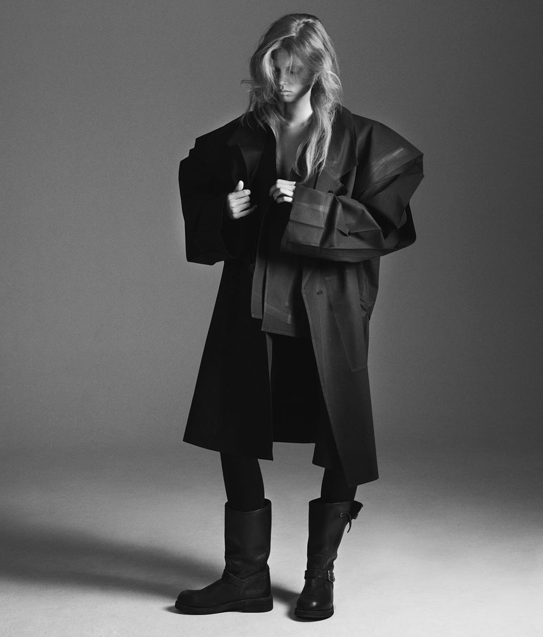Model in oversized dark coat and tall boots against a neutral studio background in a black and white portrait.