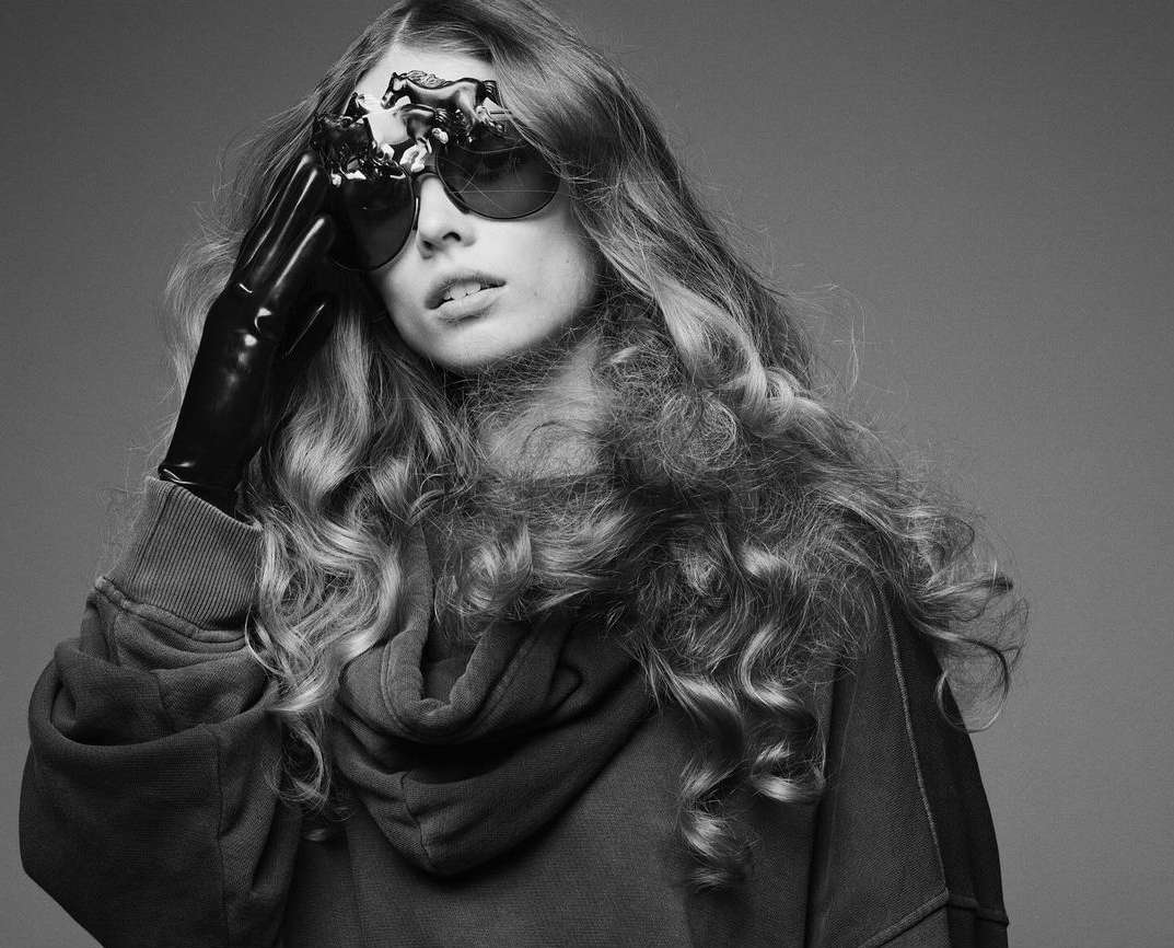 Black and white portrait of a woman wearing sunglasses with miniature horses attached, a hooded sweatshirt, and shiny black gloves.