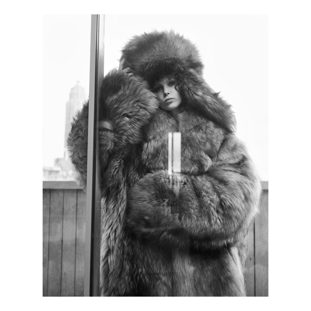Model in large fluffy fur coat and matching ushanka hat posing indoors by window with city view, photographed by Theo Sion.