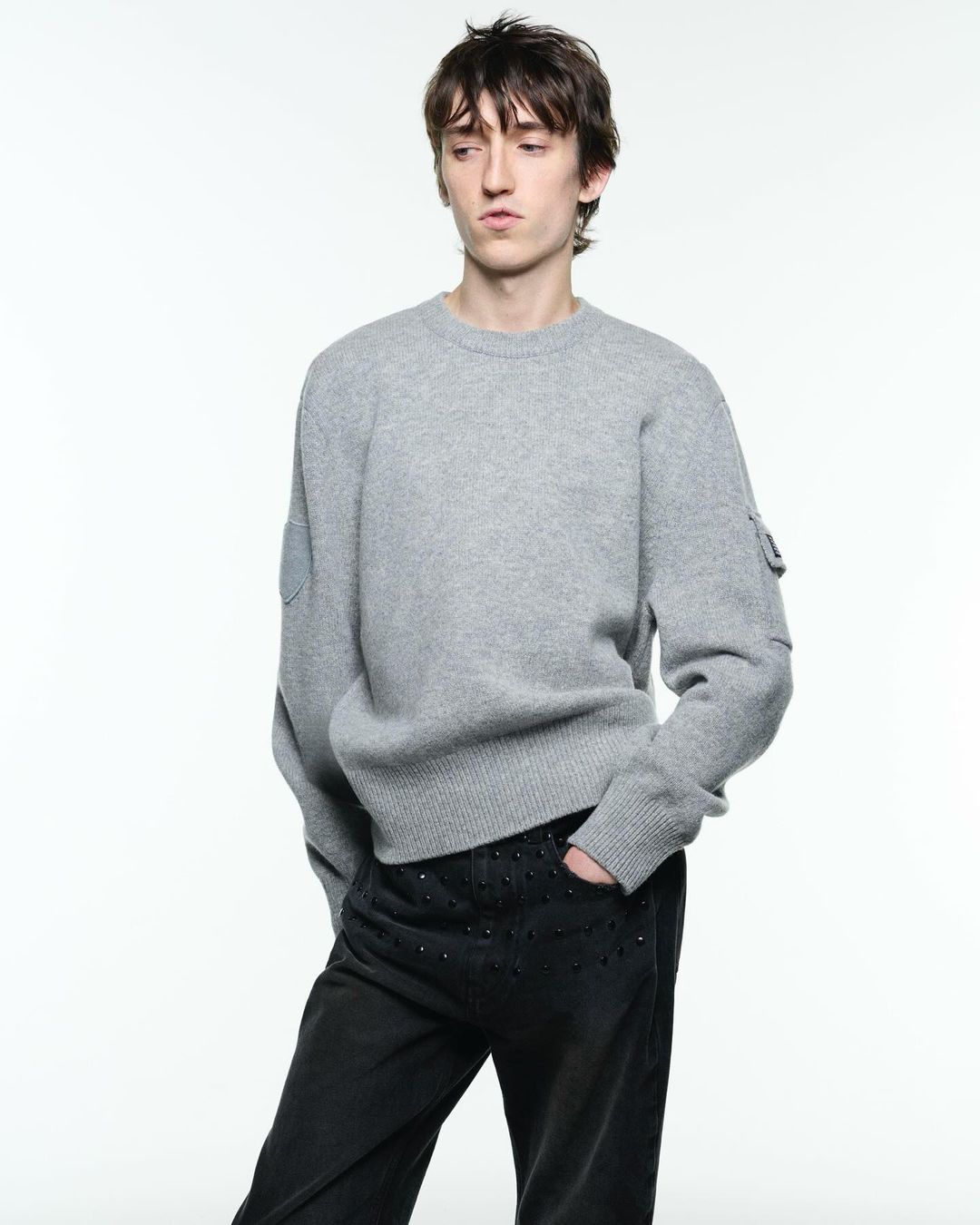 Male fashion model poses in a studio wearing a grey sweater and black pants with embellishments, looking off to the side.
