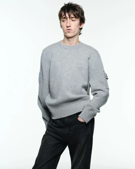 Male fashion model poses in a studio wearing a grey sweater and black pants with embellishments, looking off to the side.