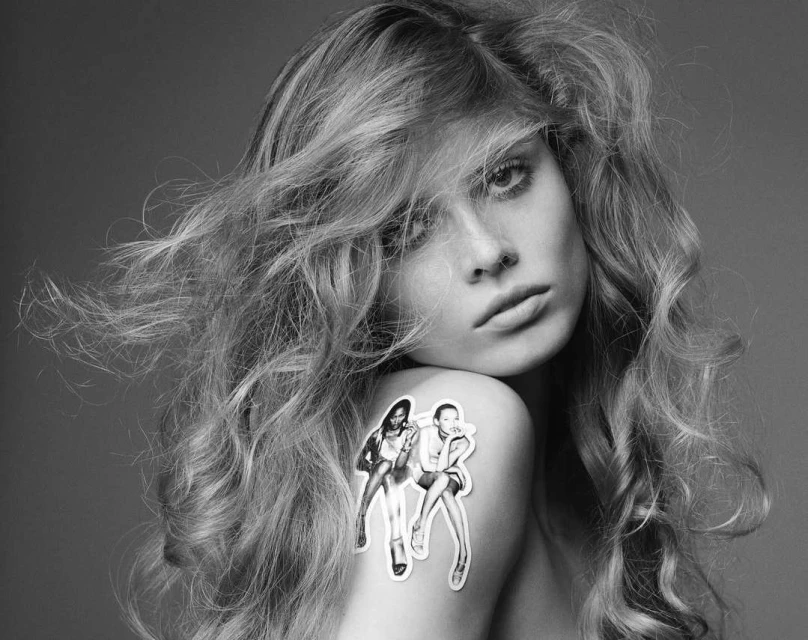 Black and white fashion portrait featuring a young fair-skinned woman with flowing hair and a sticker of two women on her shoulder.