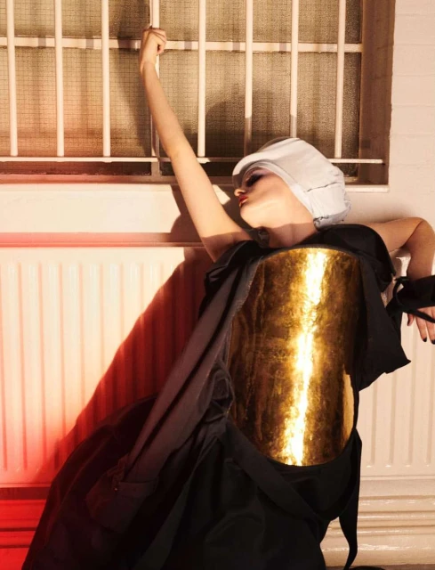 Woman in stylized dark dress with metallic front panel poses with arm raised against a barred window and pink-toned walls.