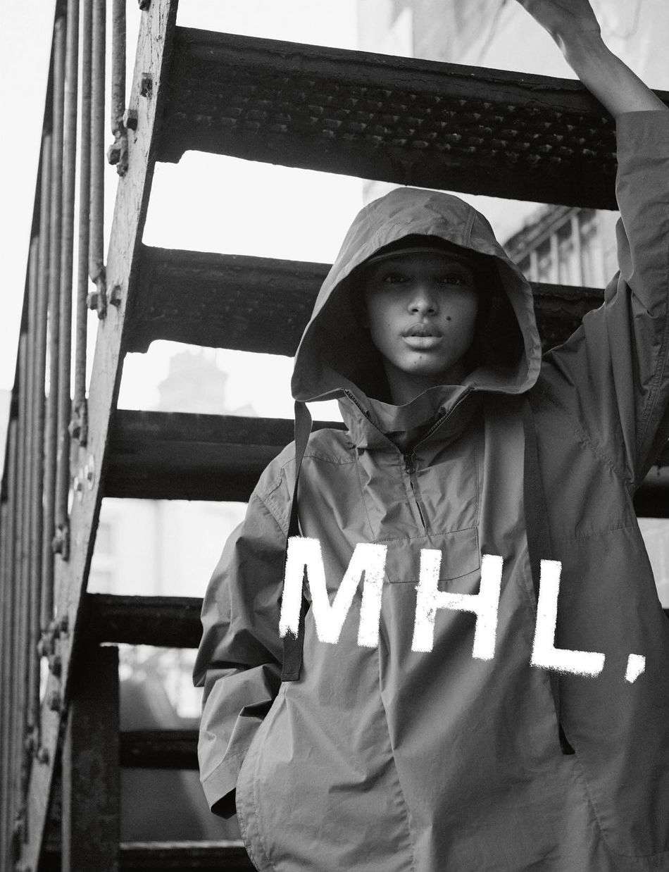 A person wearing a hooded anorak poses on metal stairs, with the MHL brand logo visible on the front of the garment in black and white.