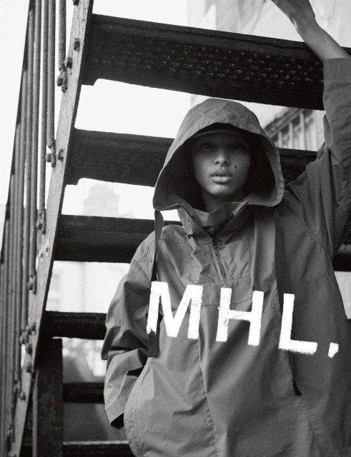 A person wearing a hooded anorak poses on metal stairs, with the MHL brand logo visible on the front of the garment in black and white.