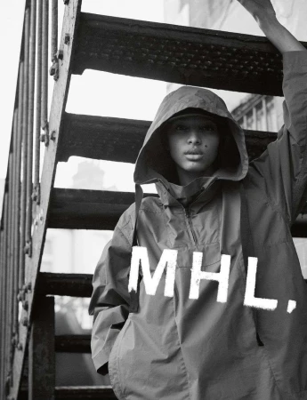 A person wearing a hooded anorak poses on metal stairs, with the MHL brand logo visible on the front of the garment in black and white.