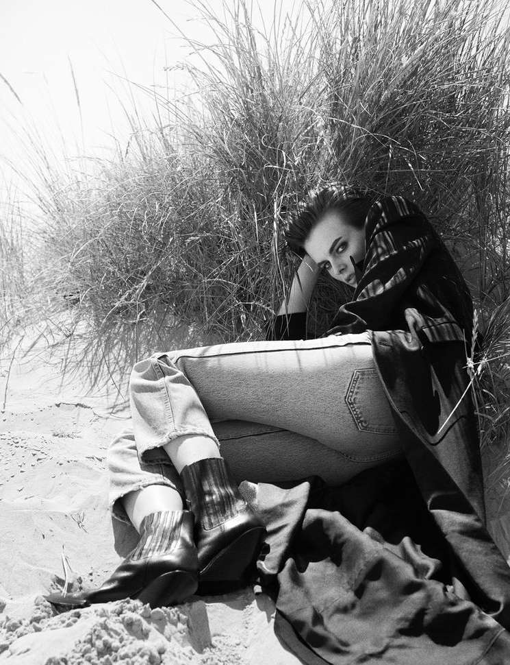 Black and white portrait of a person sitting on a beach with tall grass, wearing jeans, patterned socks and ankle boots.