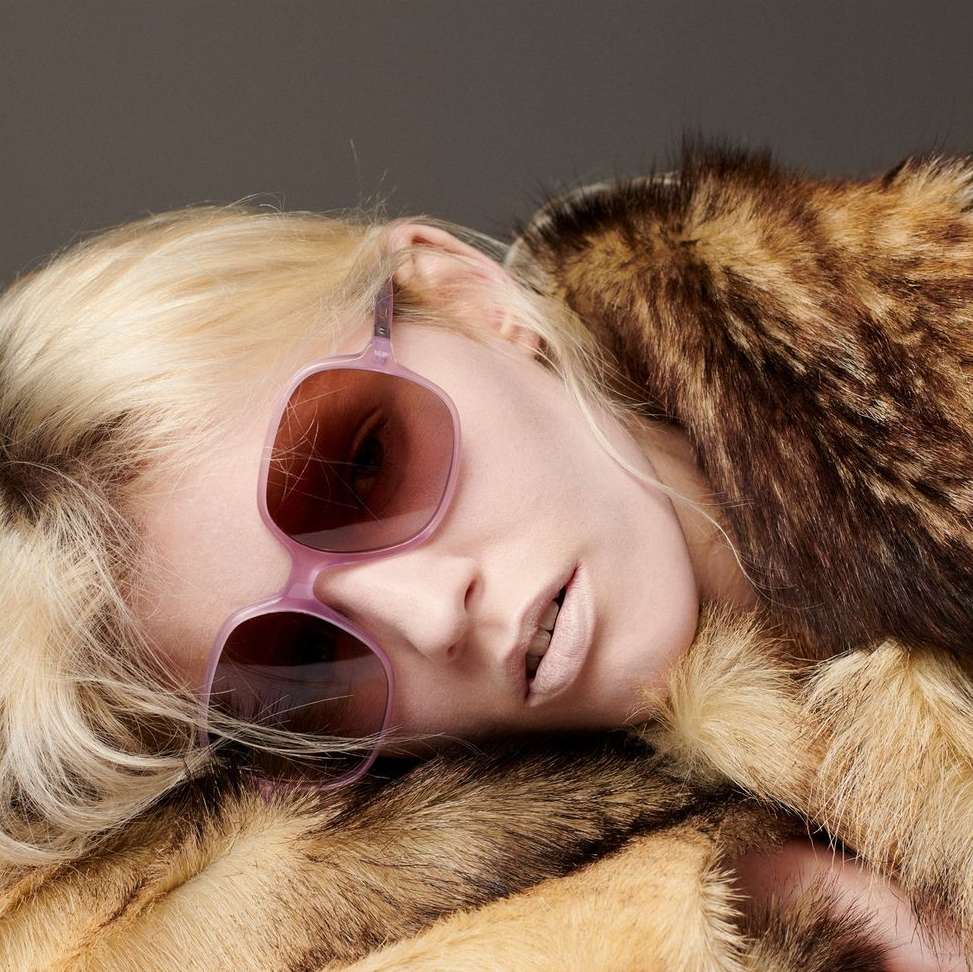Close-up of a blond woman with light skin wearing pink sunglasses and a brown fur coat against a grey background in a studio setting.