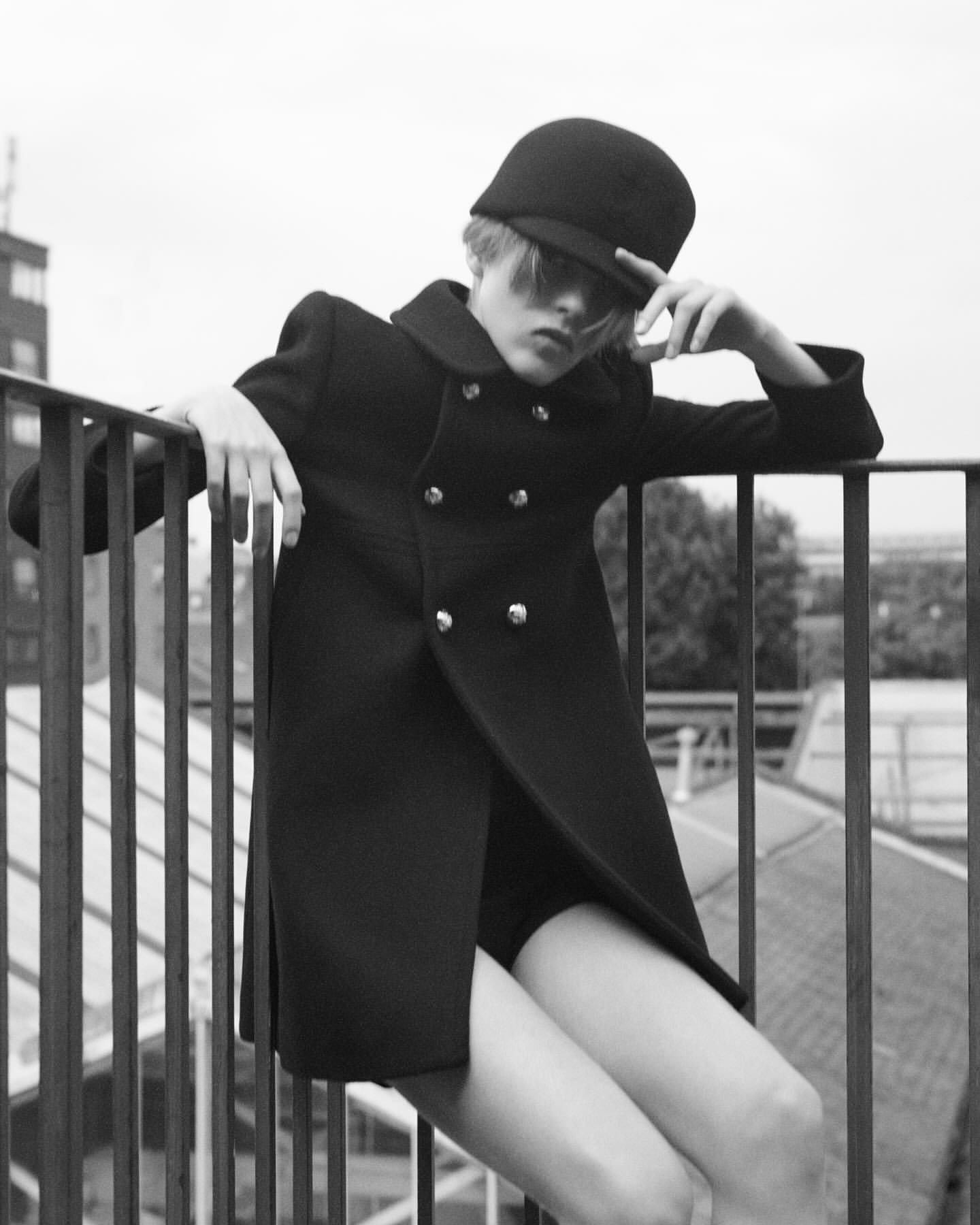 Black and white portrait of a young woman in a dark coat and cap, leaning against a railing with a cityscape in the background.