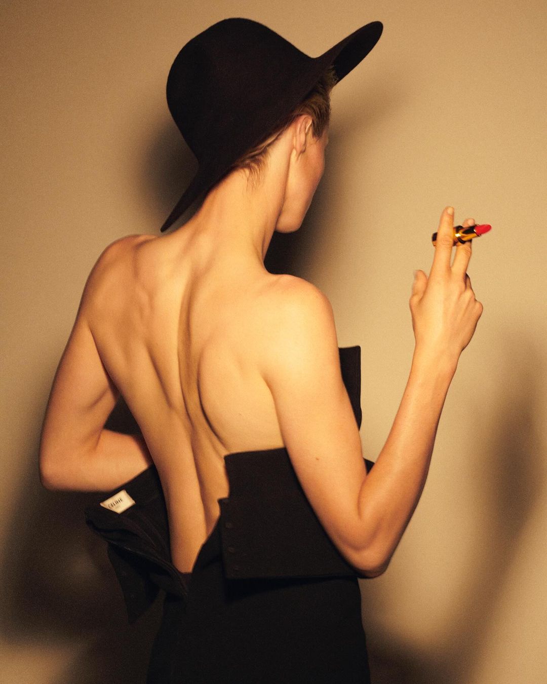 Back view of a woman wearing a black hat and partial black dress, holding a lipstick against a beige background in a studio setting.