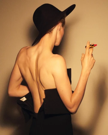 Back view of a woman wearing a black hat and partial black dress, holding a lipstick against a beige background in a studio setting.