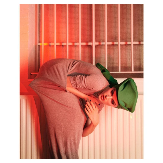 A woman in a grey dress and green hat bends, with a red-lit metal-barred window behind and a radiator visible at the bottom of the frame.