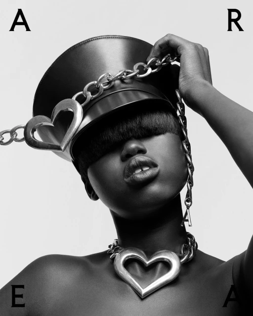 Striking black and white portrait of a woman with a unique leather hat and bold, heart-shaped chain jewelry against a light background.