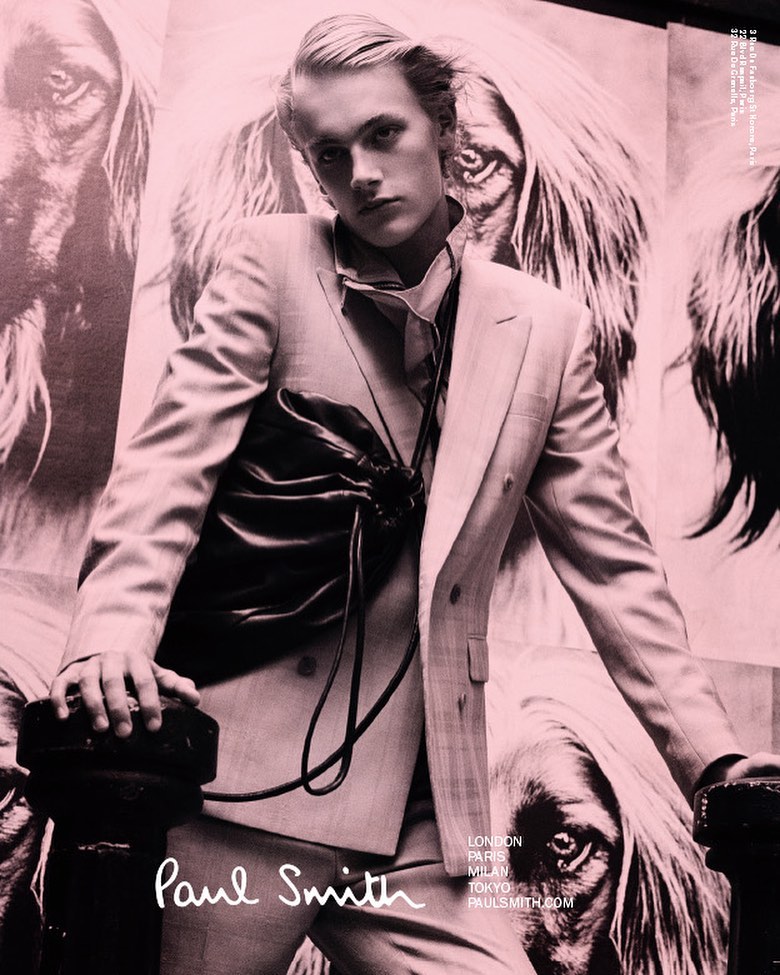 Fashion editorial shot: A blond male model in a pink suit stands in front of a dog portrait, wearing a black drawstring bag by Paul Smith.