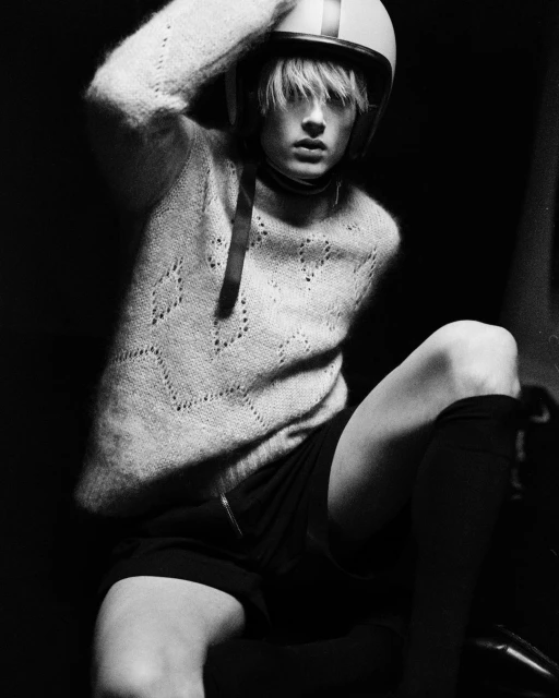 Monochrome portrait of a person with fair skin wearing a cream sweater, black shorts, and helmet indoors in a high-contrast setting.