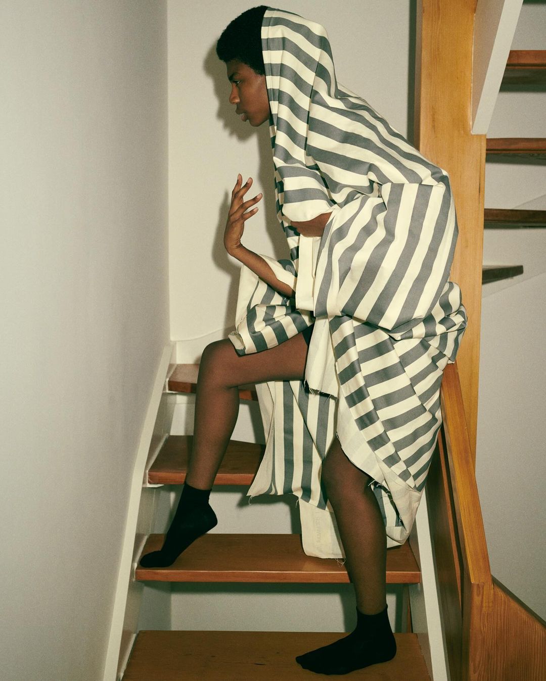African American fashion model poses on wooden stairs wearing a striped white and grey hooded robe with black socks and neutral background.