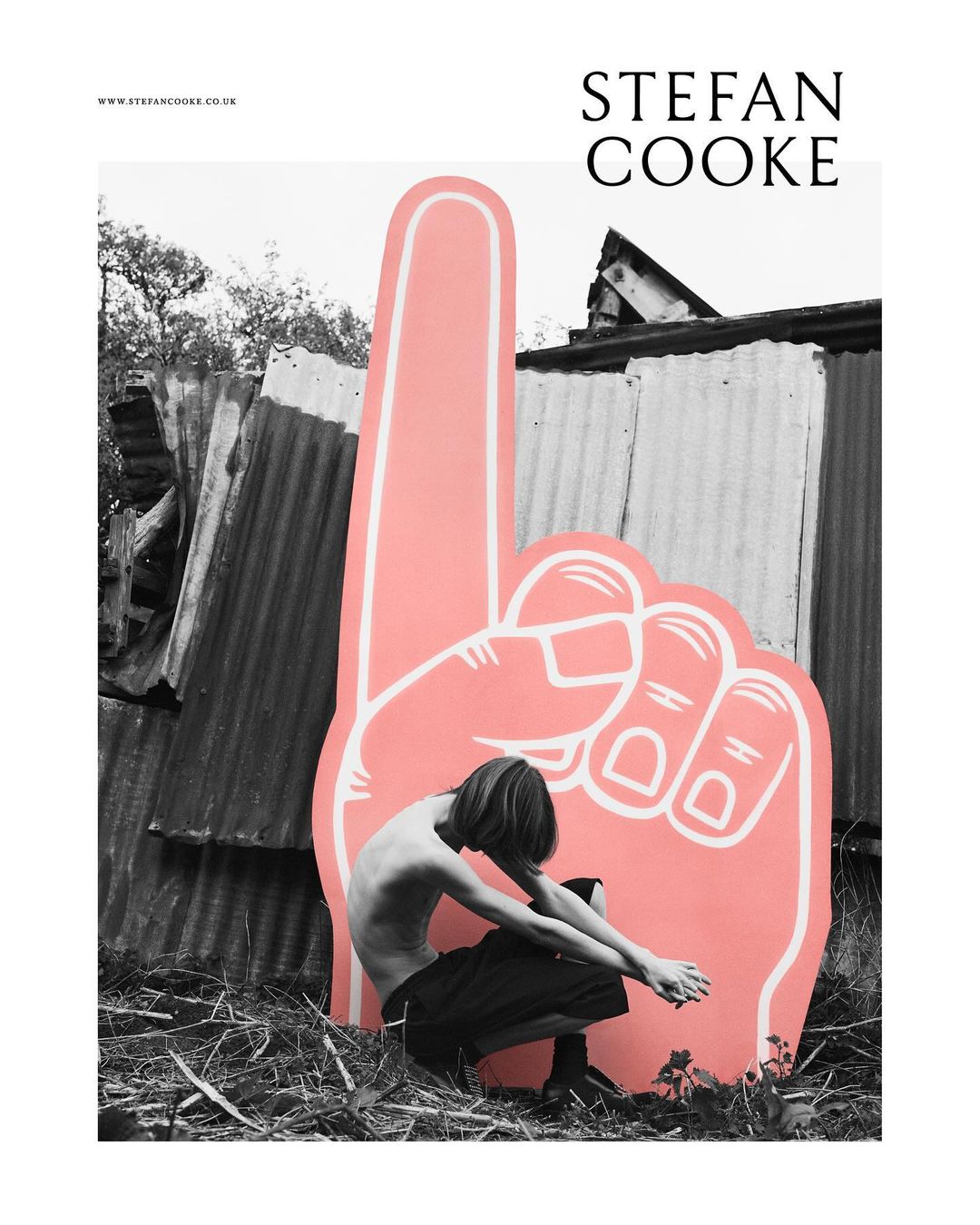 Monochrome image features a shirtless person crouching next to a large pink foam hand, set against a corrugated metal fence outdoors.