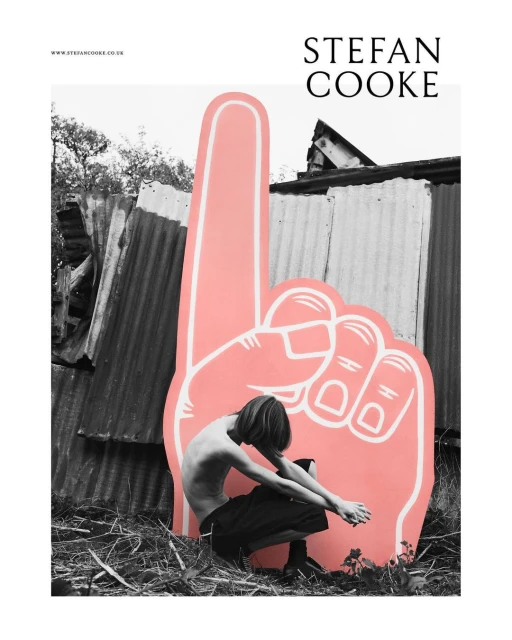 Monochrome image features a shirtless person crouching next to a large pink foam hand, set against a corrugated metal fence outdoors.