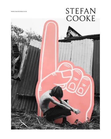 Monochrome image features a shirtless person crouching next to a large pink foam hand, set against a corrugated metal fence outdoors.