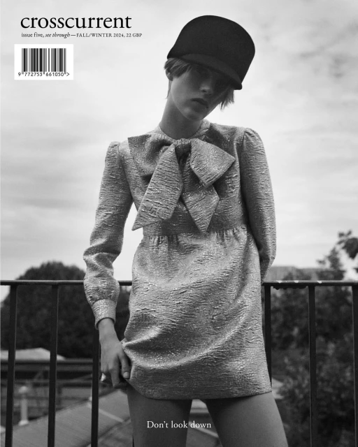 Black and white fashion shot of a slender woman wearing a bow-tie dress and black hat standing outdoors on a balcony overlooking trees.
