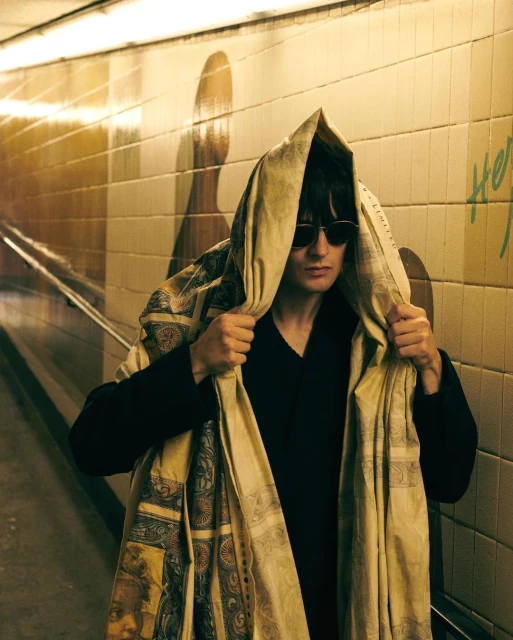 Man in sunglasses stands in tiled tunnel wearing patterned hooded wrap. Neutral color palette. Graffiti on the wall in the background.