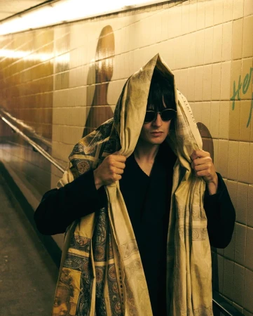 Man in sunglasses stands in tiled tunnel wearing patterned hooded wrap. Neutral color palette. Graffiti on the wall in the background.