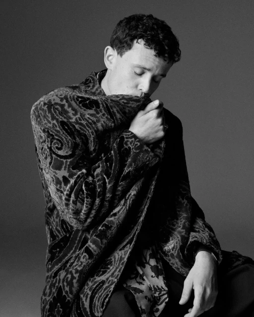 Black and white portrait of a young adult man with curly hair wearing a dark patterned velvet jacket indoors, with eyes closed.