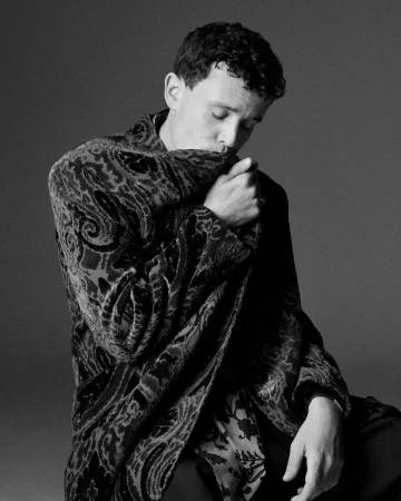 Black and white portrait of a young adult man with curly hair wearing a dark patterned velvet jacket indoors, with eyes closed.