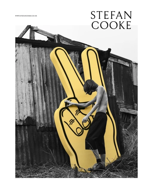 Teen poses with large yellow foam hand in front of a weathered building outdoors. Youthful fashion, black and white photography.