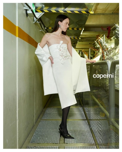 A woman poses in a white dress with a matching coat in an industrial tunnel with yellow stripes and metal grated floor for Coperni fashion.