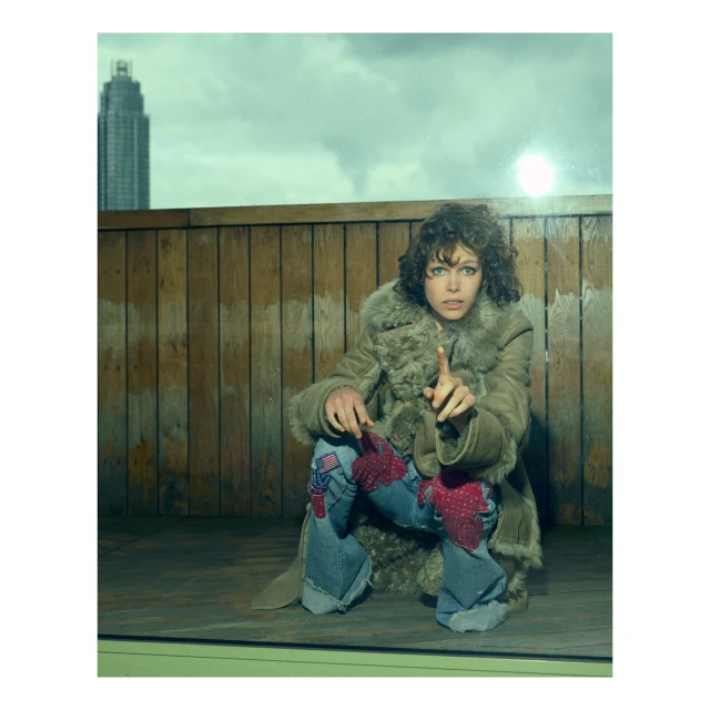 An adult female with curly brown hair kneels while pointing in a brown faux-fur coat and patchwork jeans. A building is in the background.